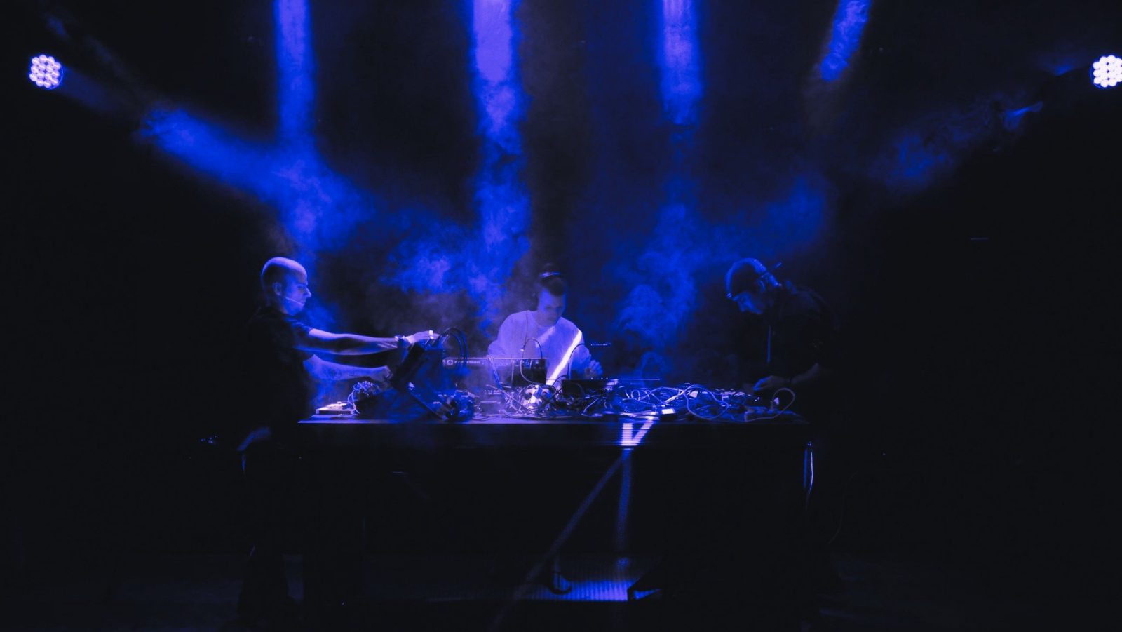 Three artists performing a live hardware set on a dark stage in blue light and haze